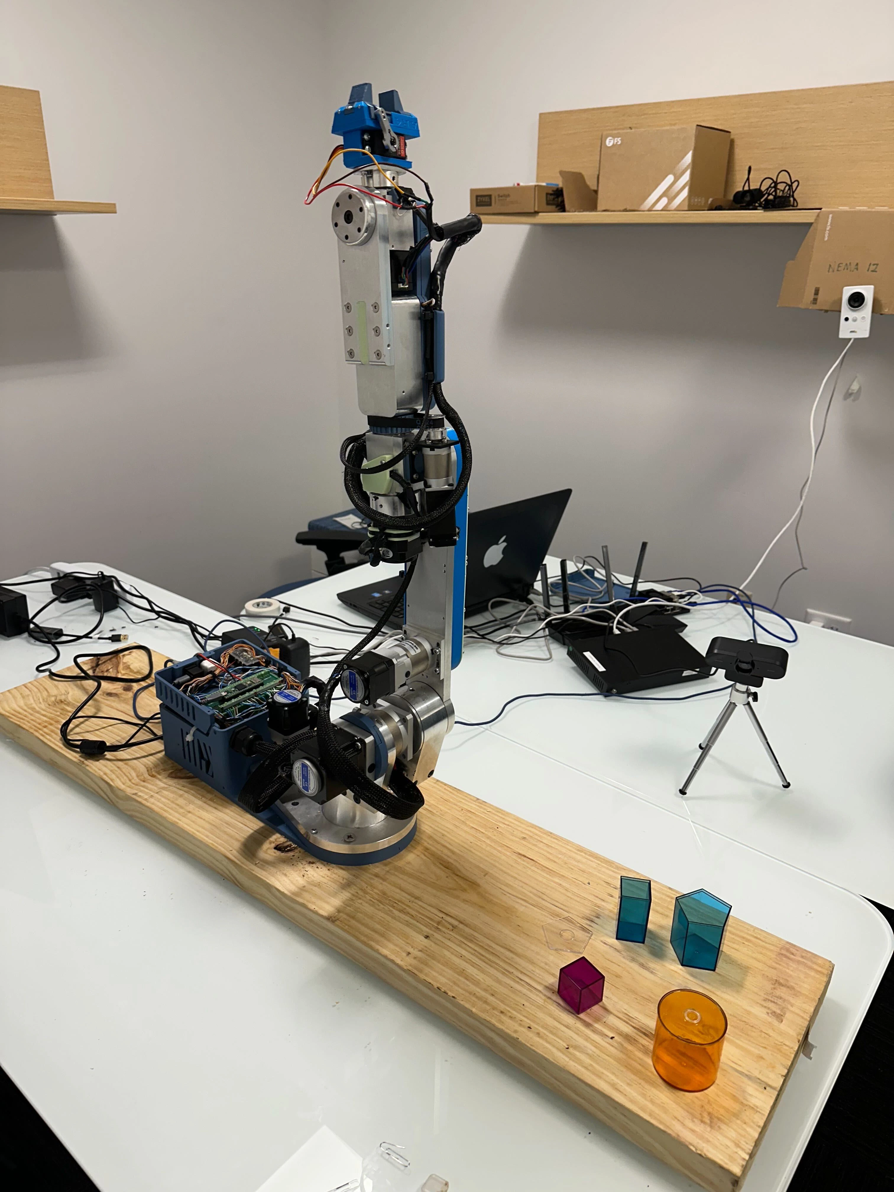 Robotic arm on a wooden board reaching toward colored geometric objects on a table, with a stereo camera head mounted above and a tripod-mounted webcam observing the scene.