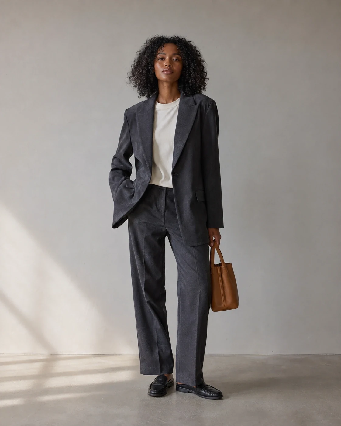 Woman in a gray suit holding a brown handbag in a pale studio