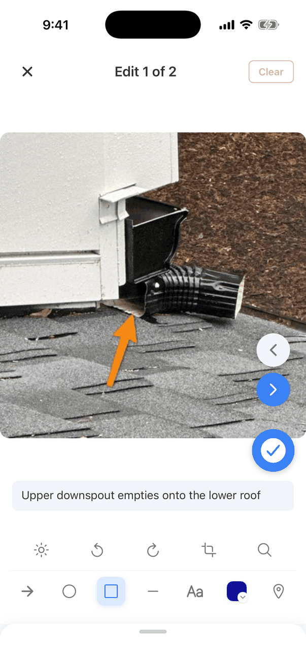 The annotation screen, titled Edit 1 of 2, with an X button and a greyed-out Clear button. The photo of a downspout fills the middle, with left and right arrow buttons and a blue tick button. Below it the caption Upper downspout empties onto the lower roof, and two rows of tools: brightness, rotate left, rotate right, crop and loupe; then arrow, circle, box (selected), line, Aa text, a blue colour swatch and a location pin