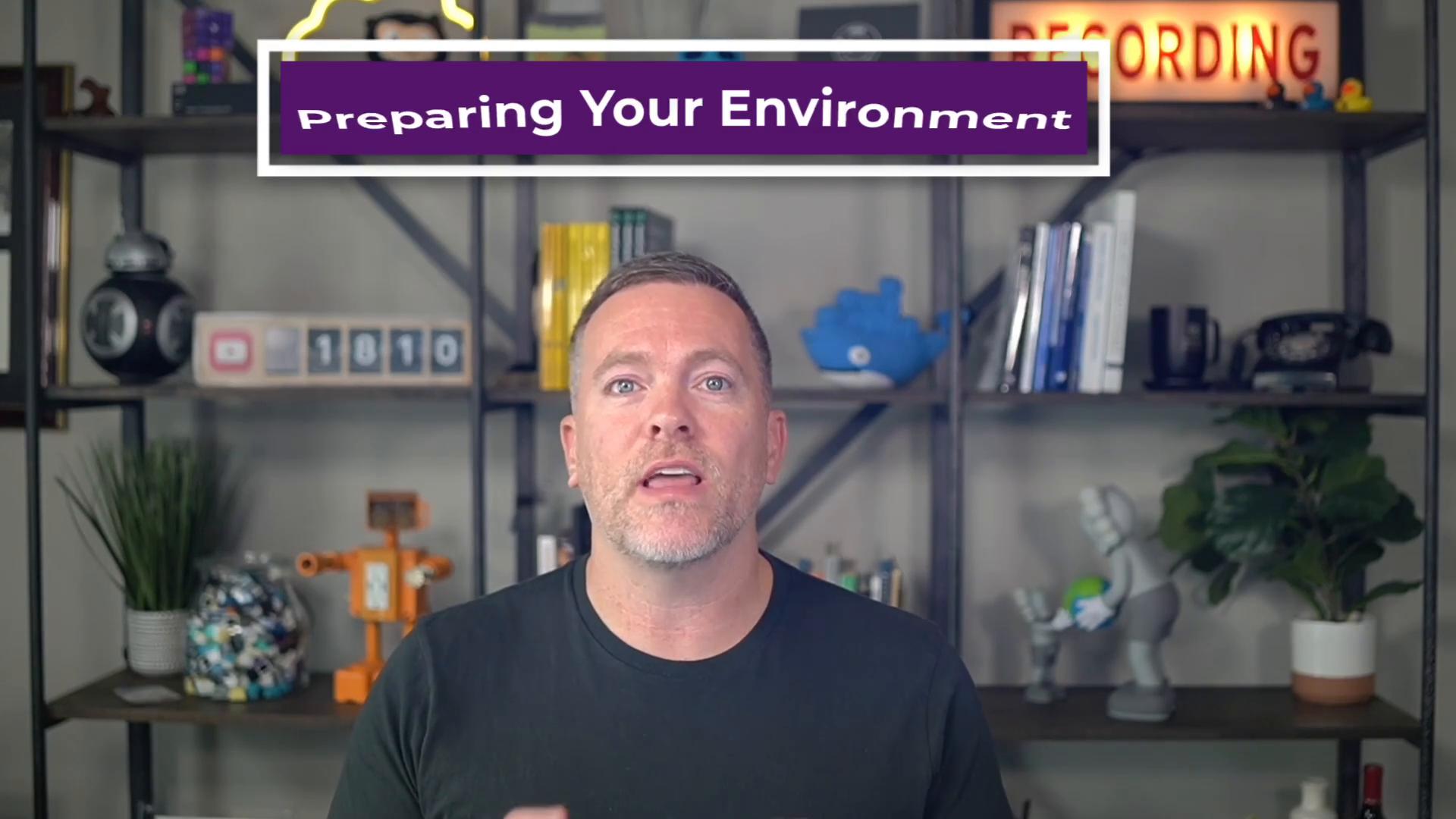 A man is speaking on camera with a purple banner above him that reads "Preparing Your Environment." Behind him is a shelving unit holding books, plants, and assorted decorative items, giving an office or studio feel.