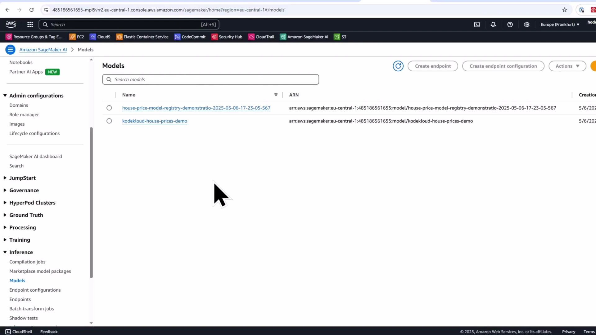 A screenshot of the Amazon SageMaker "Models" console in a web browser showing two model entries (house-price-model-registry-demonstratio... and kodekloud-house-prices-demo) with the left navigation menu visible. A large black mouse cursor is shown near the center of the screen.