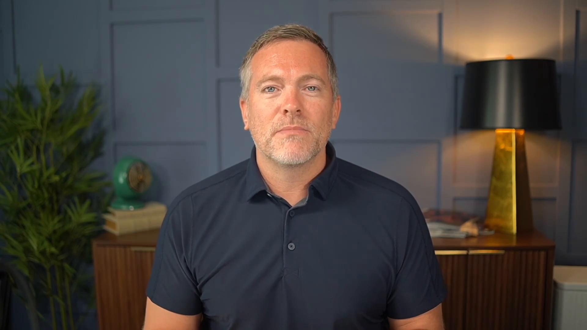 A man with short hair and stubble wearing a dark polo sits facing the camera in a neatly decorated room. Behind him are a potted plant, a small green fan on stacked books, and a gold-based table lamp against a paneled blue wall.