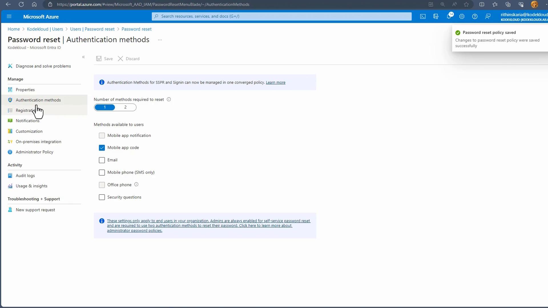 A Microsoft Azure portal screenshot showing the "Password reset | Authentication methods" settings, with the number of methods required set to 1 and "Mobile app code" checked. The left navigation menu and a top-right notification saying "Password reset policy saved" are also visible.