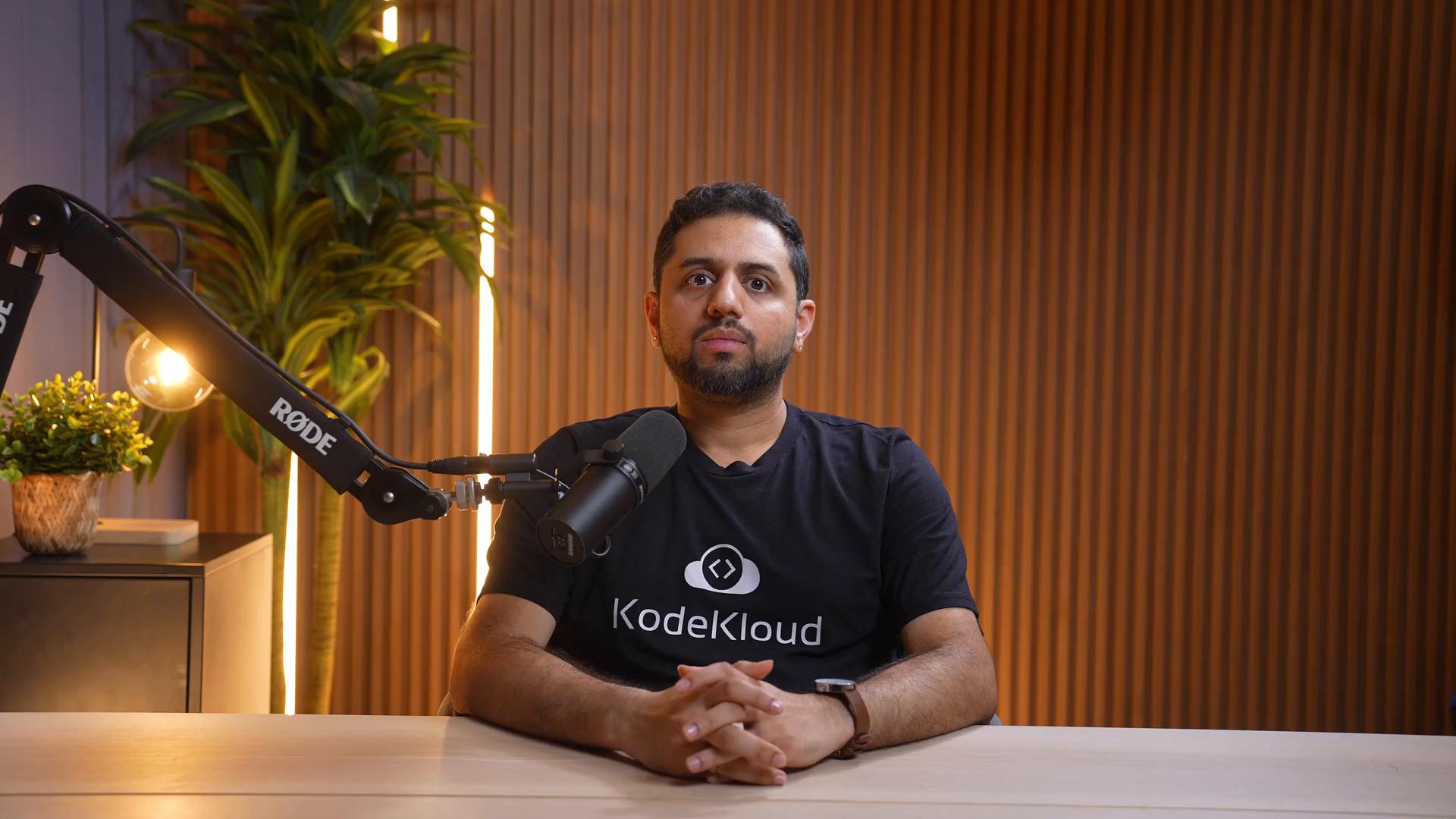 A man sits at a desk in a recording studio, facing the camera with a microphone on a boom arm in front of him. The background features a potted plant, warm vertical wood paneling, and soft lighting.