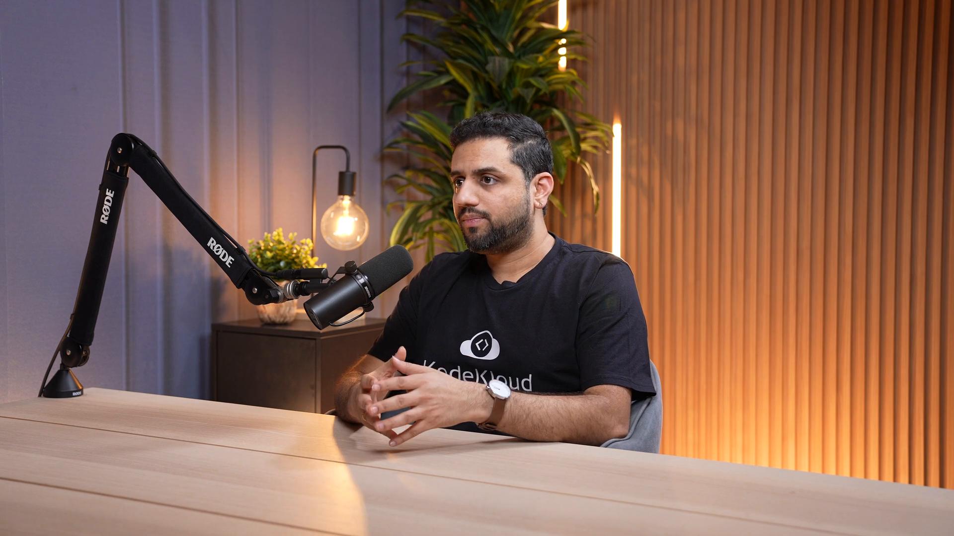 A man sits at a table speaking into a Rode microphone on a boom arm in a warmly lit podcast/studio setup with plants and a lamp behind him. He gestures with his hands as he talks.