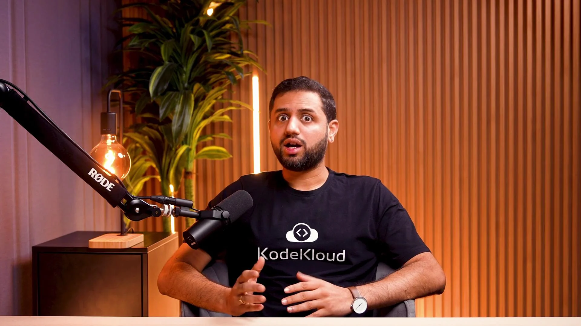 A bearded man wearing a KodeKloud t‑shirt sits in front of a microphone with a surprised expression, gesturing with his hands. He’s in a warm, well-lit recording setup with a lamp, plant, and wooden slatted wall behind him.