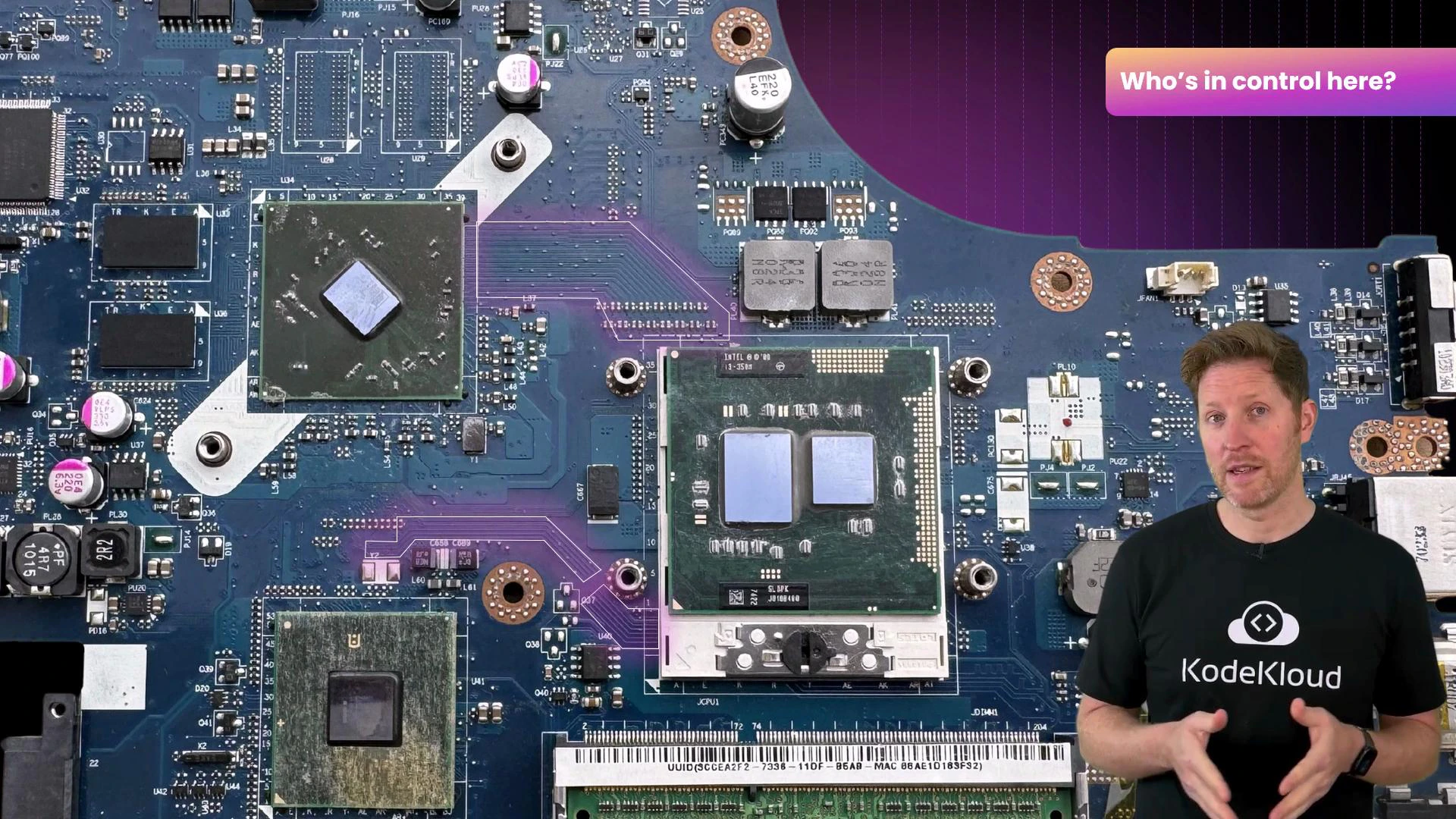 A close-up of a computer motherboard showing exposed chips, sockets, and electronic components. A man wearing a black KodeKloud T-shirt stands to the right, gesturing in front of the board.