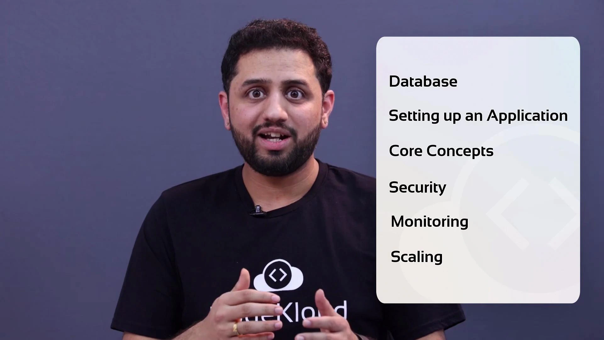 A bearded man in a black T-shirt speaks into a lapel microphone against a plain purple-gray background. A sidebar lists topics: Database, Setting up an Application, Core Concepts, Security, Monitoring, and Scaling.