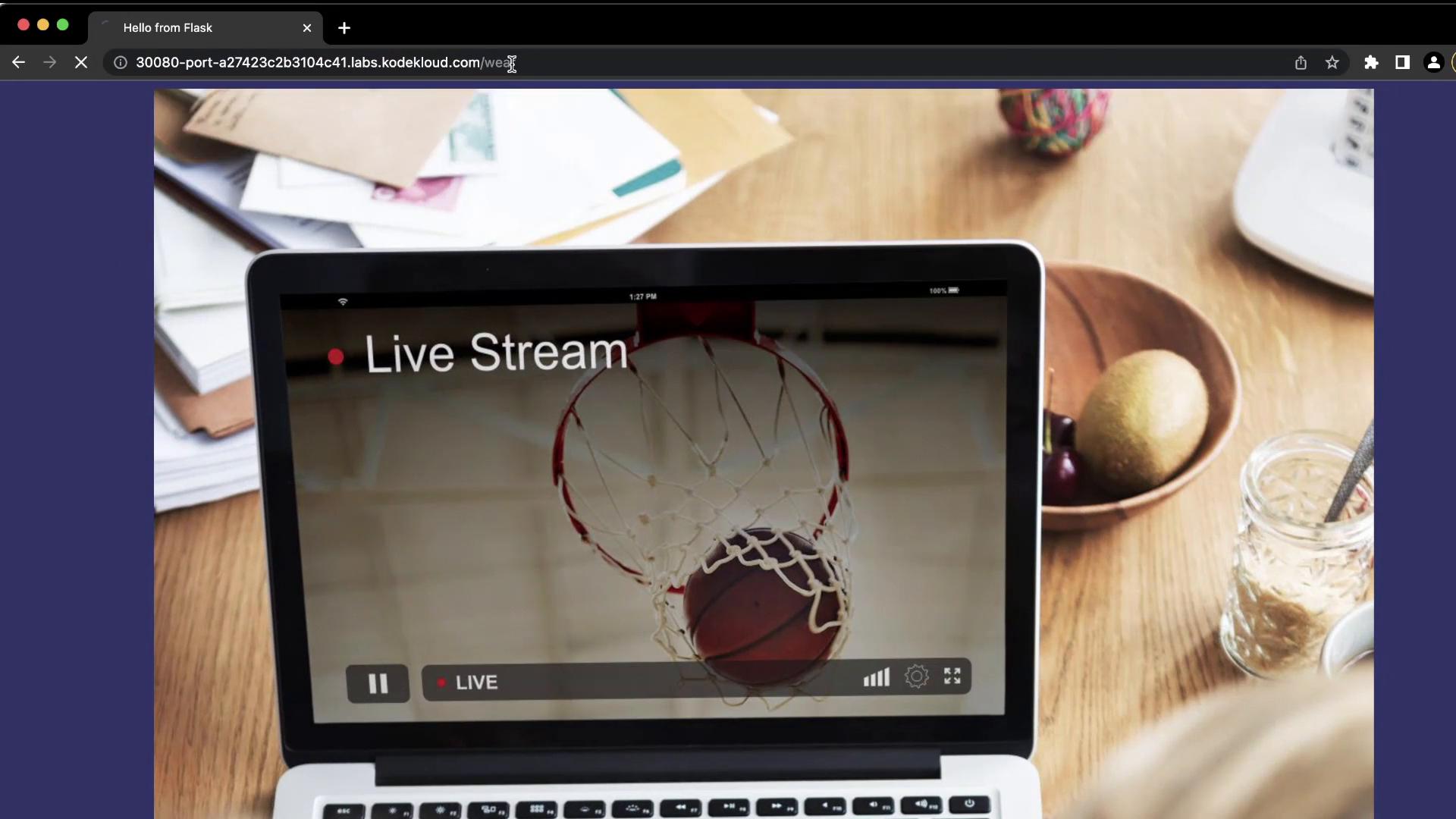 A laptop on a wooden desk displaying a live-stream video of a basketball going through a hoop with "Live Stream" shown on the screen. Papers, a fruit bowl and a glass jar sit nearby on the table.