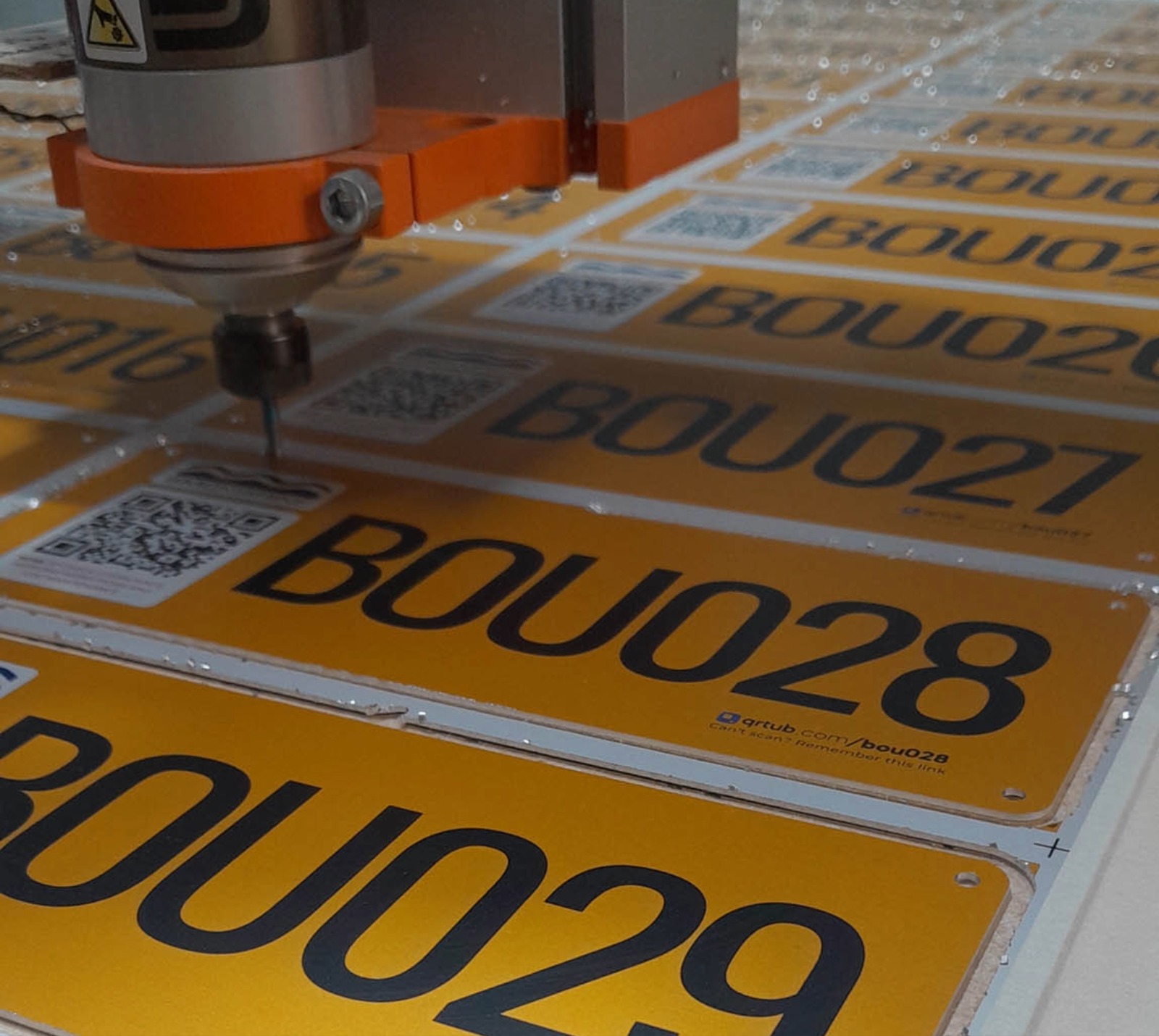 A CNC machine cutting a sheet of photo-anodized aluminum asset plates, each carrying a QR code, a large asset number and a readable link. The visible plates run in sequence: BOU027, BOU028, BOU029.