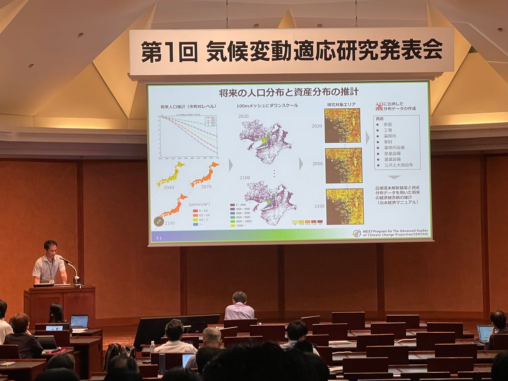 A speaker at a lectern beneath the meeting banner, presenting maps and charts of projected population and asset distributions to the audience.