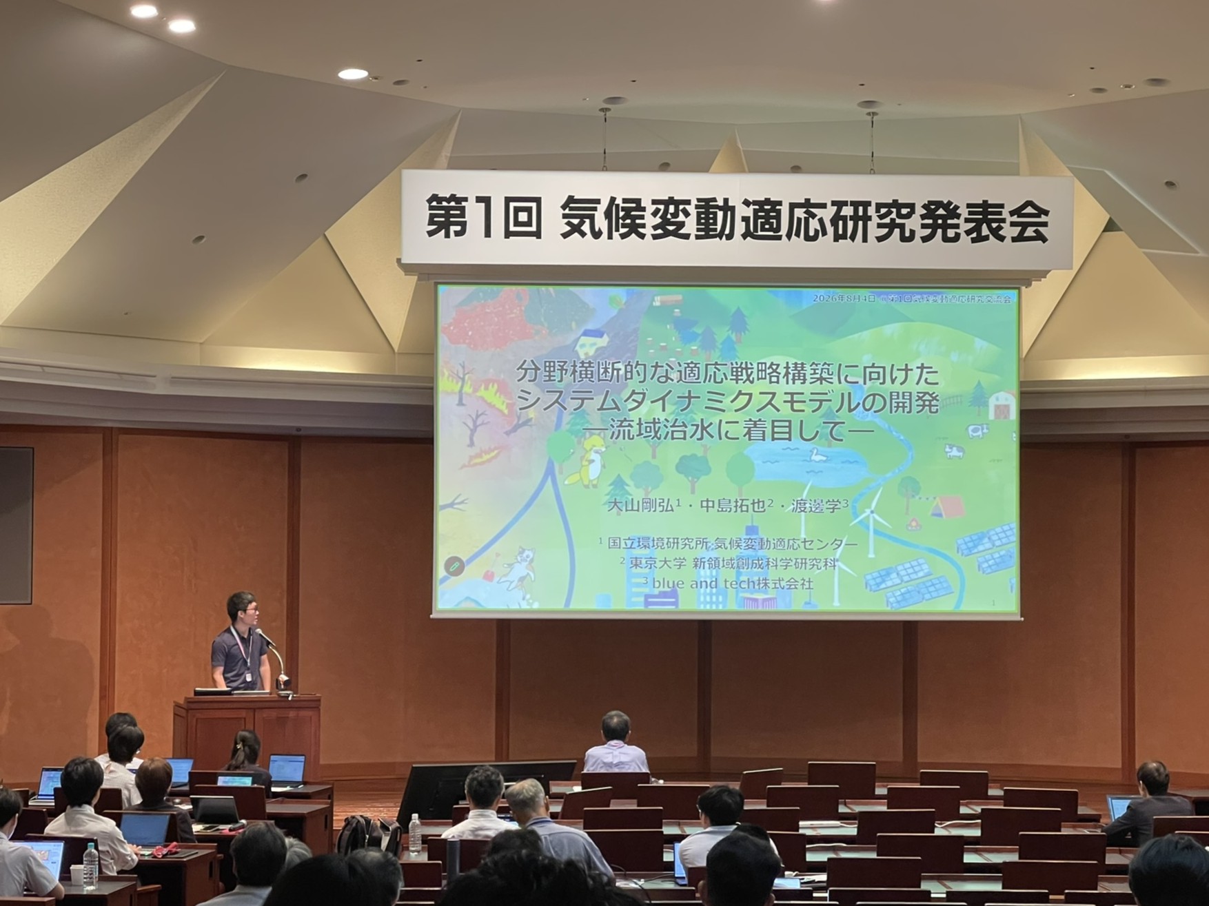 An auditorium session with a speaker beside a projected title slide about system dynamics modelling for adaptation strategies, illustrated with a river catchment.