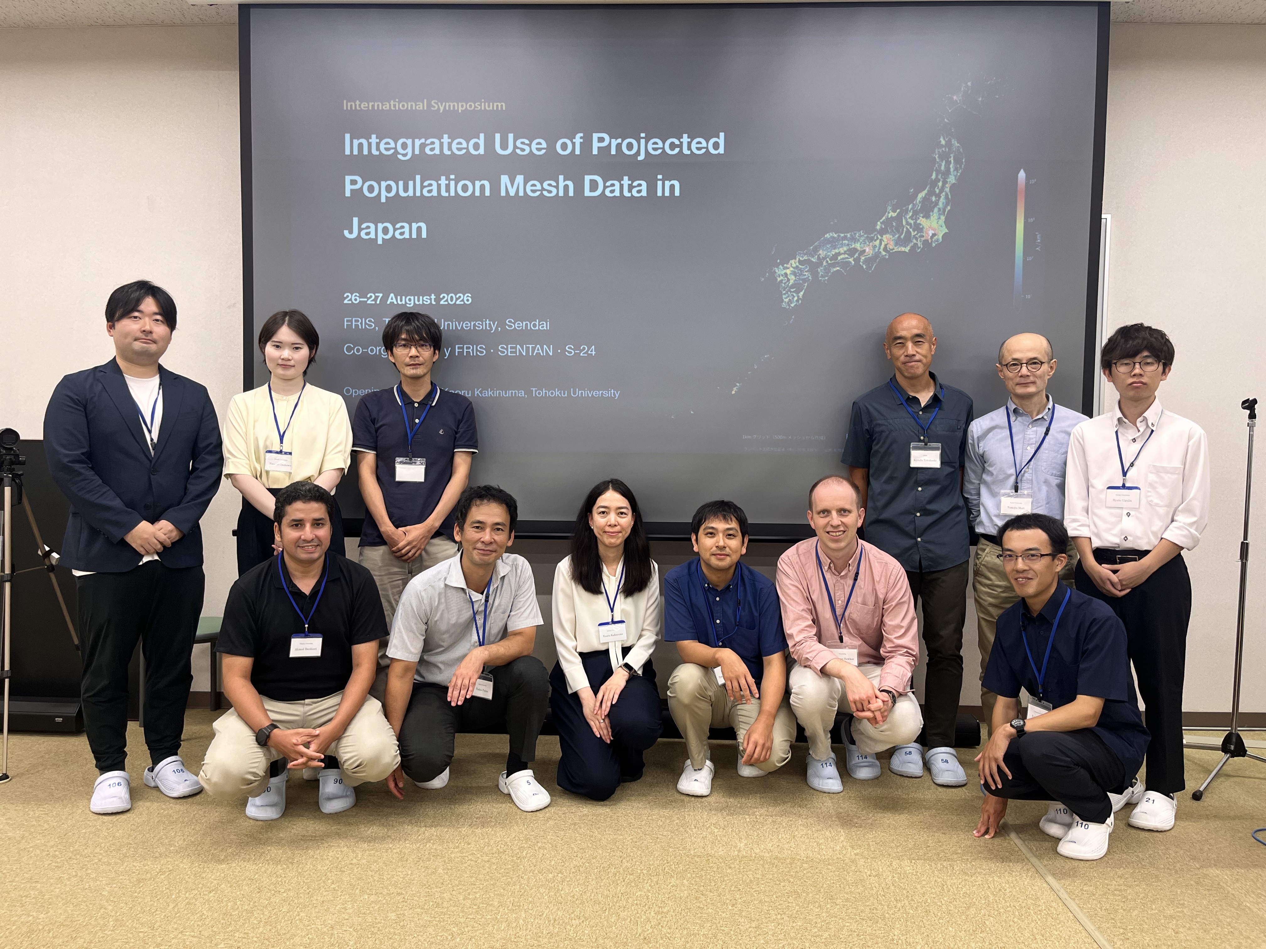 Symposium participants gathered in front of a screen displaying the event title and a population mesh map of Japan.
