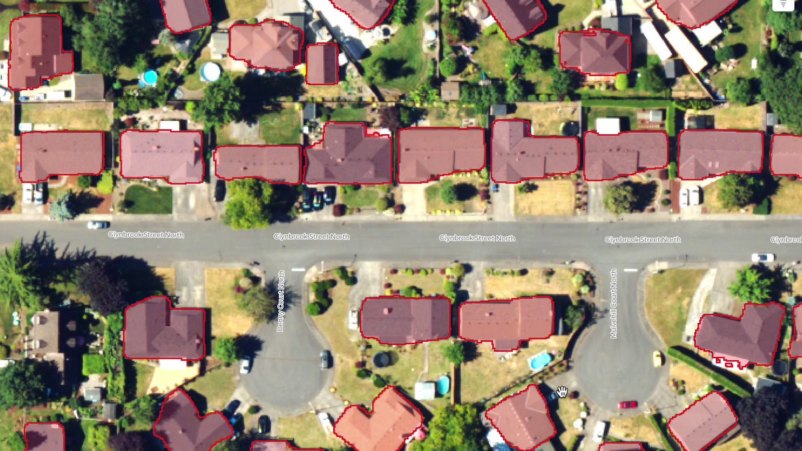 Close-up of 30 cm aerial imagery over a residential street, with red polygons outlining each house roof. The polygons follow complex L-shaped and T-shaped rooflines closely. Several small outbuildings and a detached garage carry no polygon.