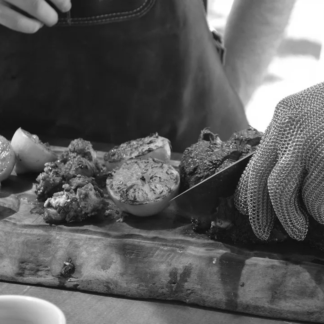 Primer plano en blanco y negro de una mano usando un guante de malla metálica mientras corta carne asada junto a vegetales a la parrilla.