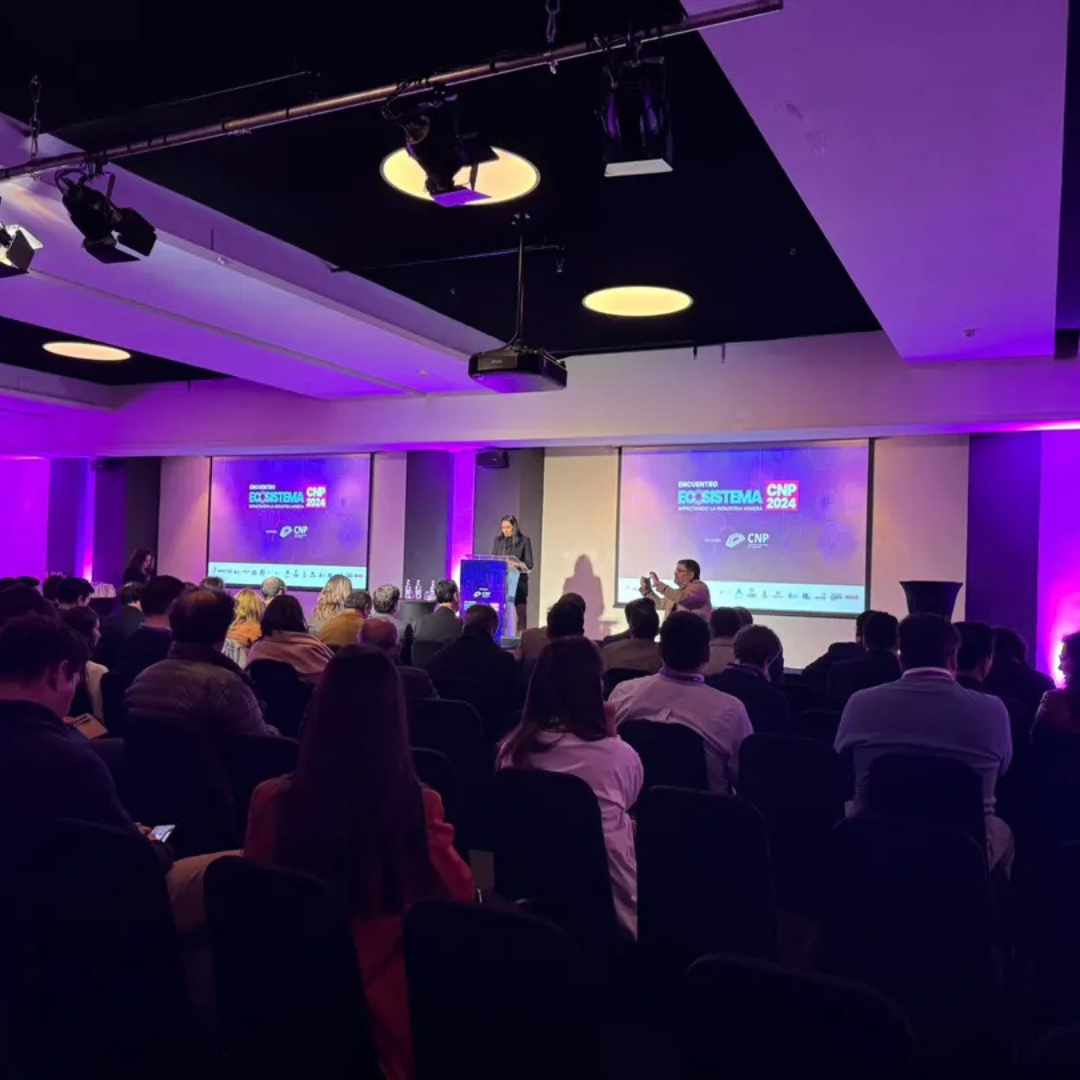 Audiencia y presentador en el escenario durante el evento 'Encuentro Ecosistema CNP 2024', con pantallas iluminadas y una atmósfera de iluminación púrpura.