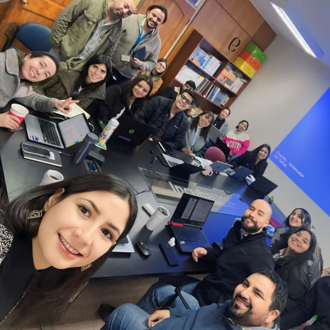 Grupo de personas sonriendo y posando para una selfie en una sala de reuniones, con laptops y documentos en la mesa.